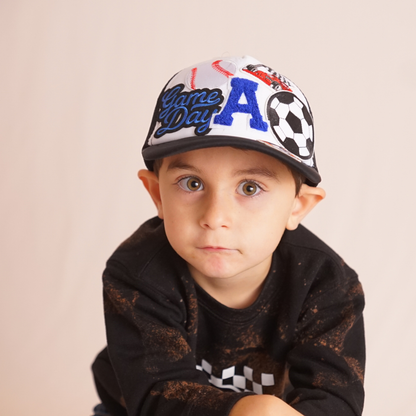 Child wearing a colorful cap with 'Game Day' text and a soccer ball design, against a plain background.