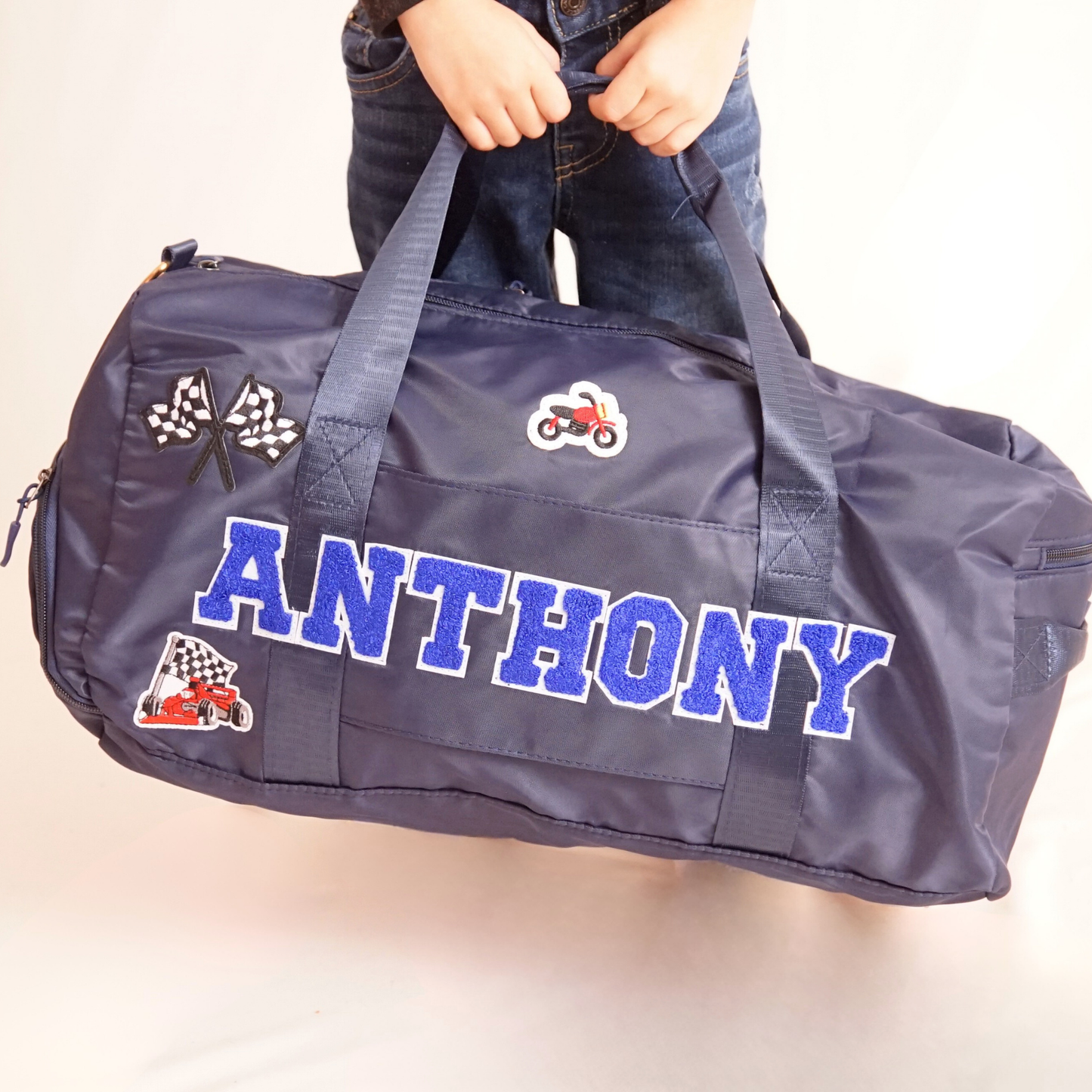 Child holding navy blue duffle bag with name ANTHONY and various patches.