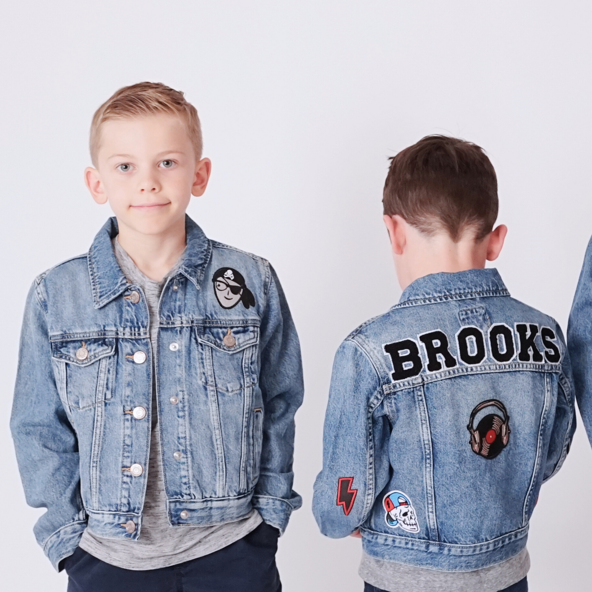 Two children wearing denim jackets with patches and text, one facing forward and the other back.