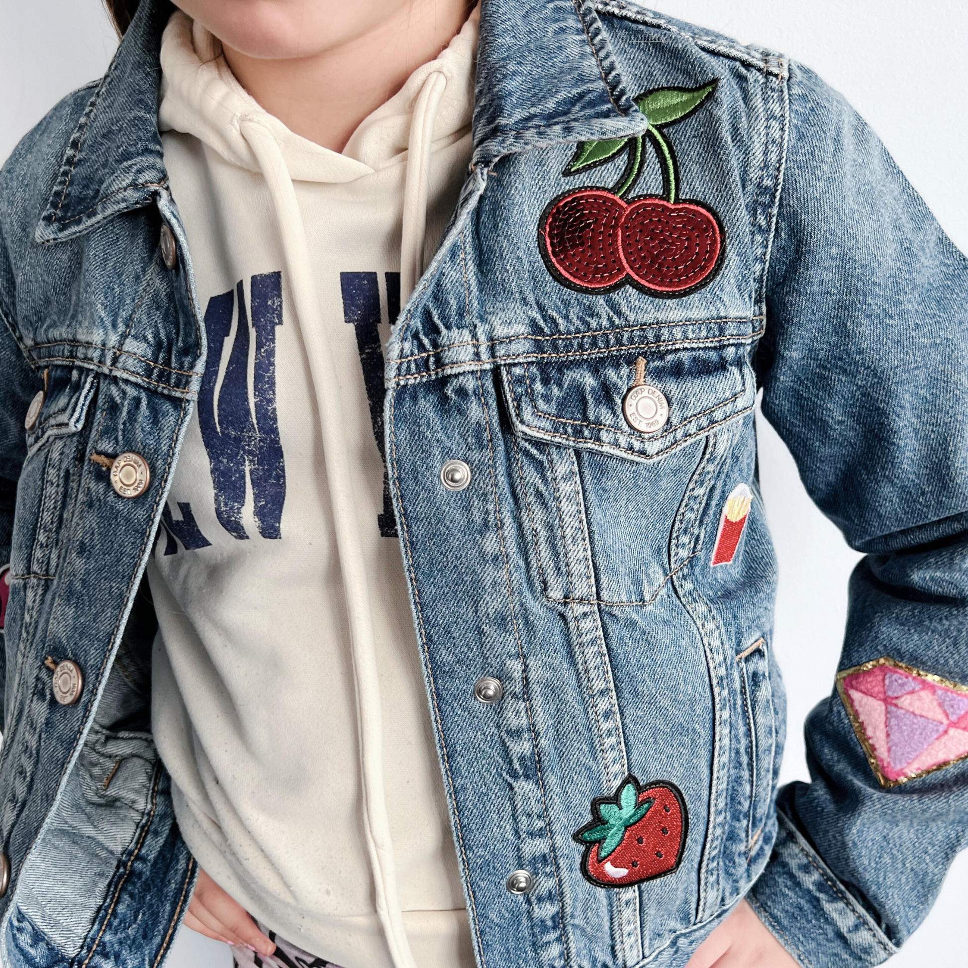 Person wearing a denim jacket with colorful patches including cherries and strawberries.