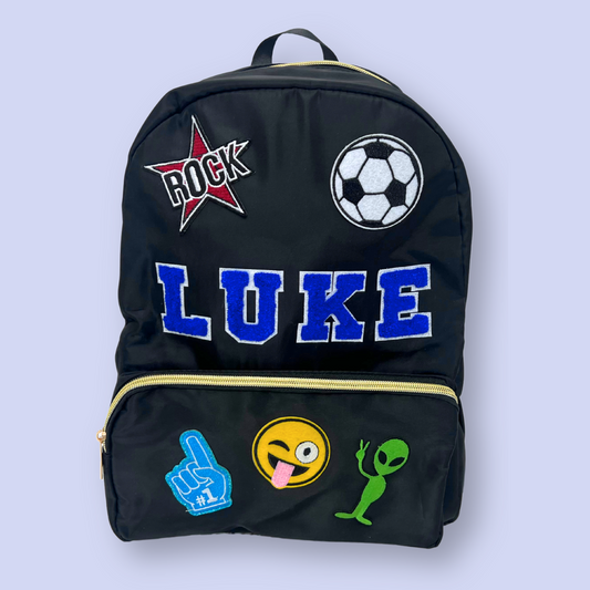 Black backpack with various patches including a star, soccer ball, and emojis on a light blue background.