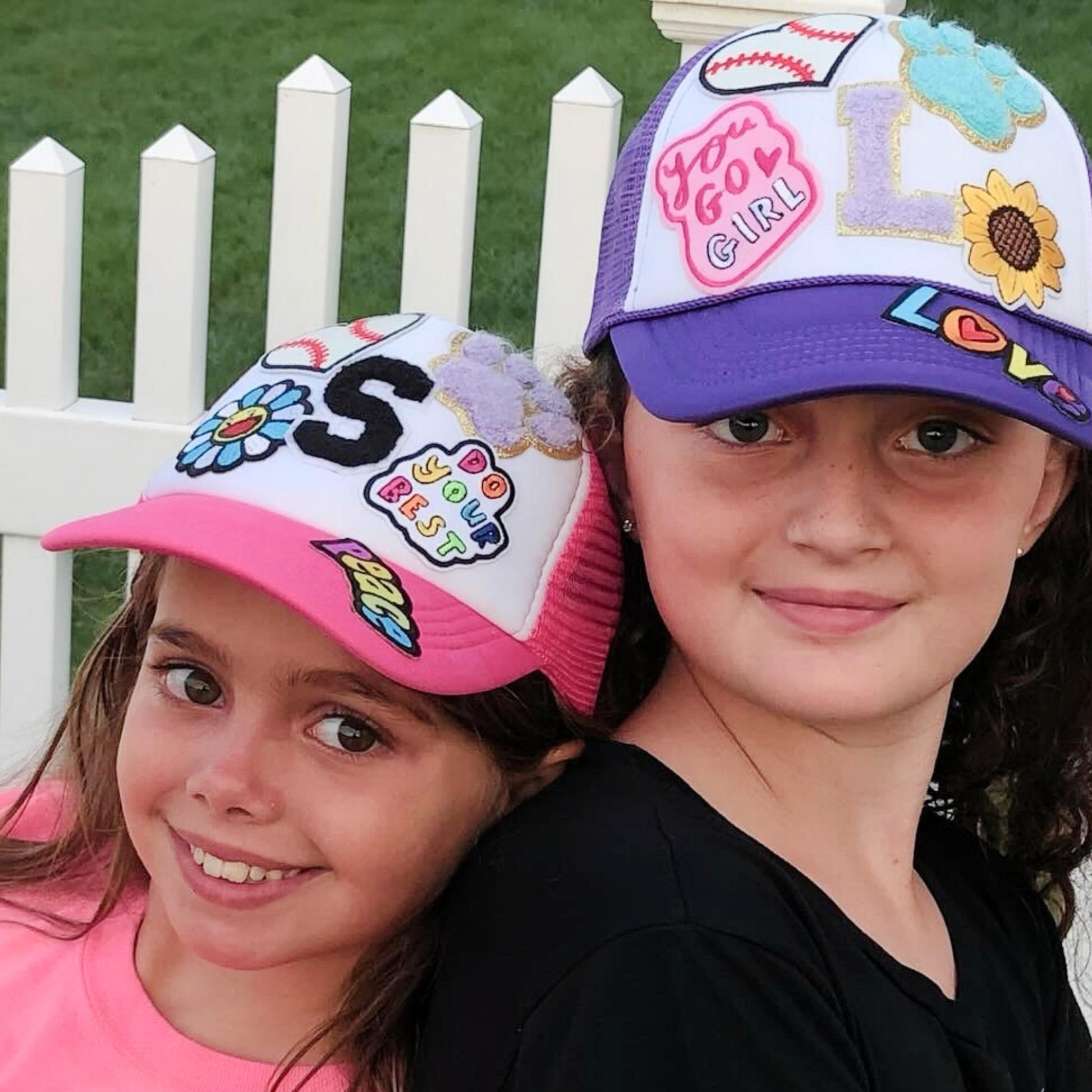 Two children wearing colorful baseball caps with decorative text and designs, standing in front of a white picket fence.