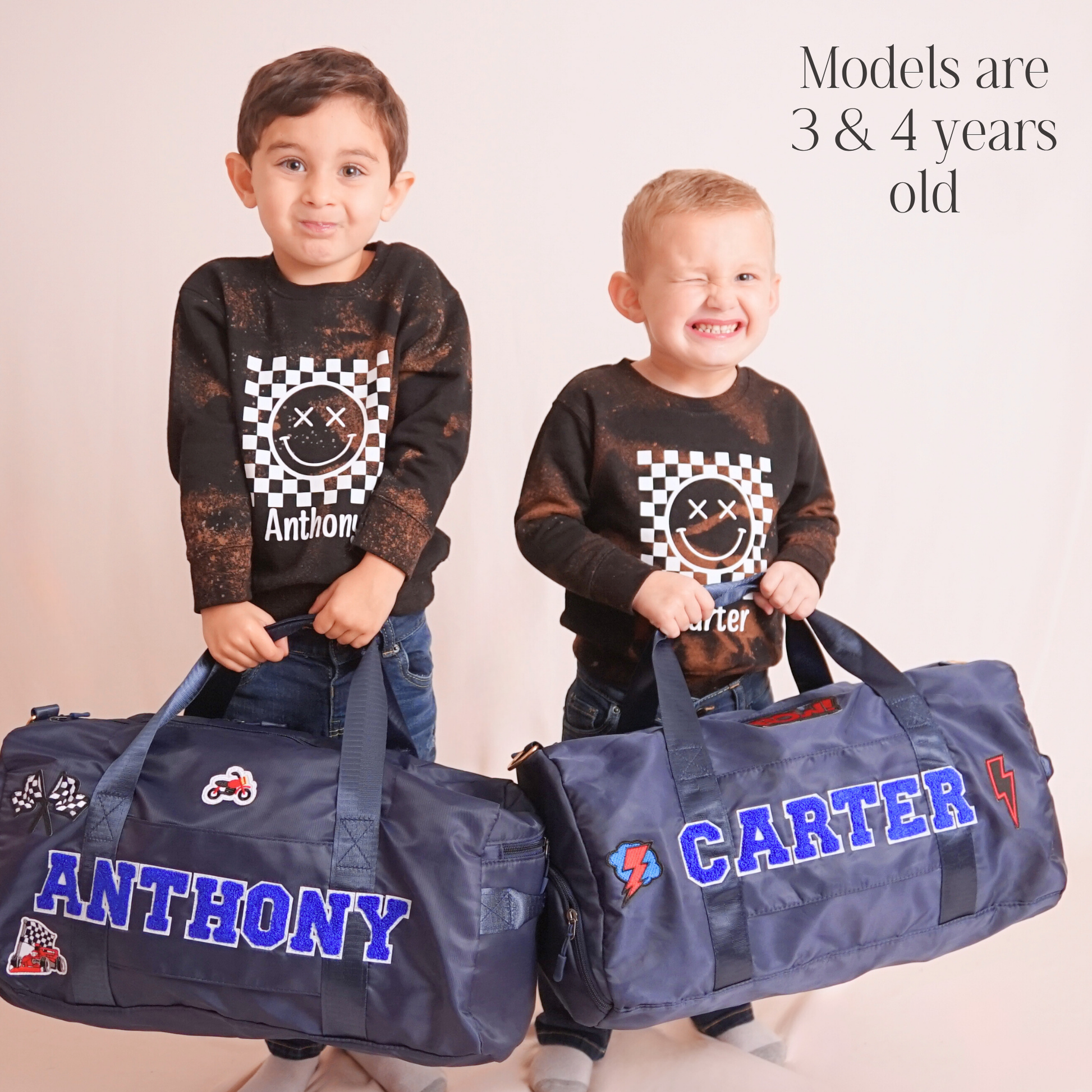 Two young boys holding navy blue duffle bags with names ANTHONY and CARTER.