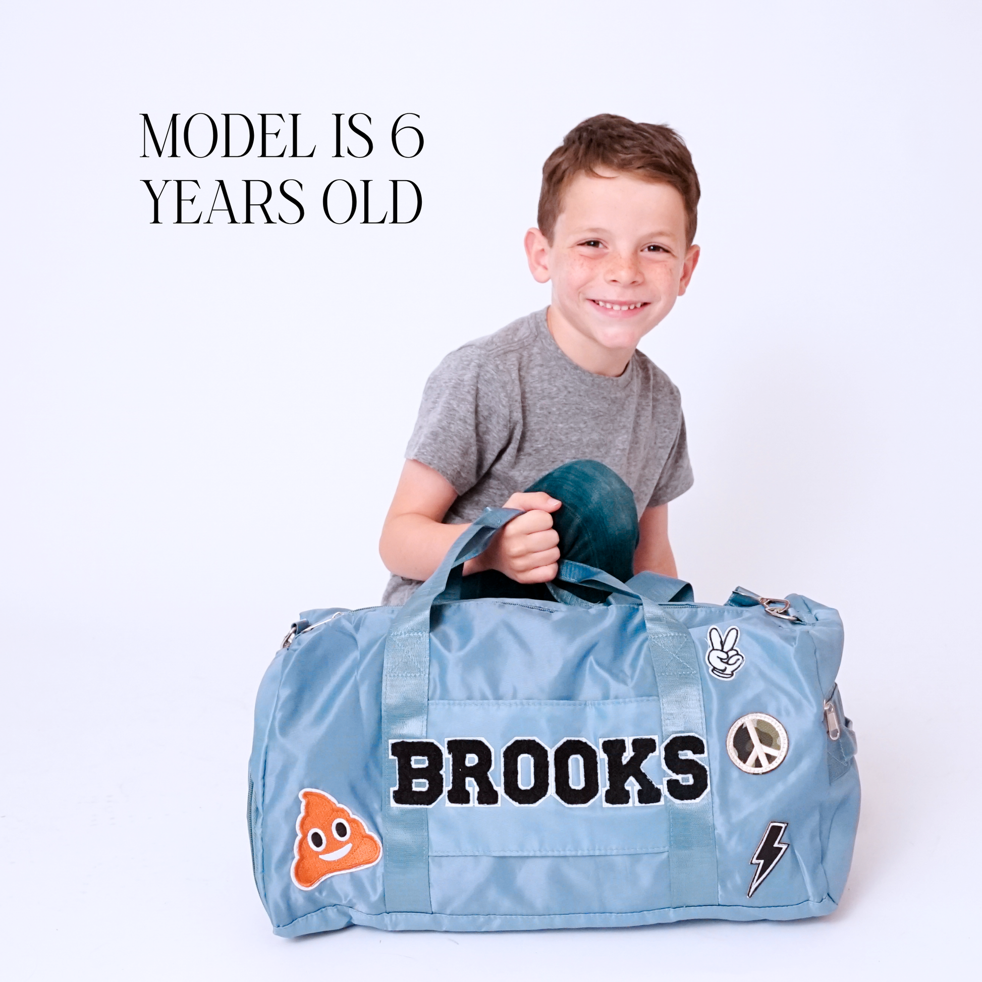 Child holding light blue duffle bag with name BROOKS and sports-themed patches.