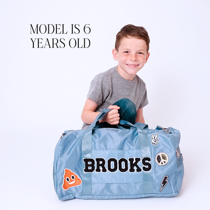 Child holding light blue duffle bag with name BROOKS and sports-themed patches.