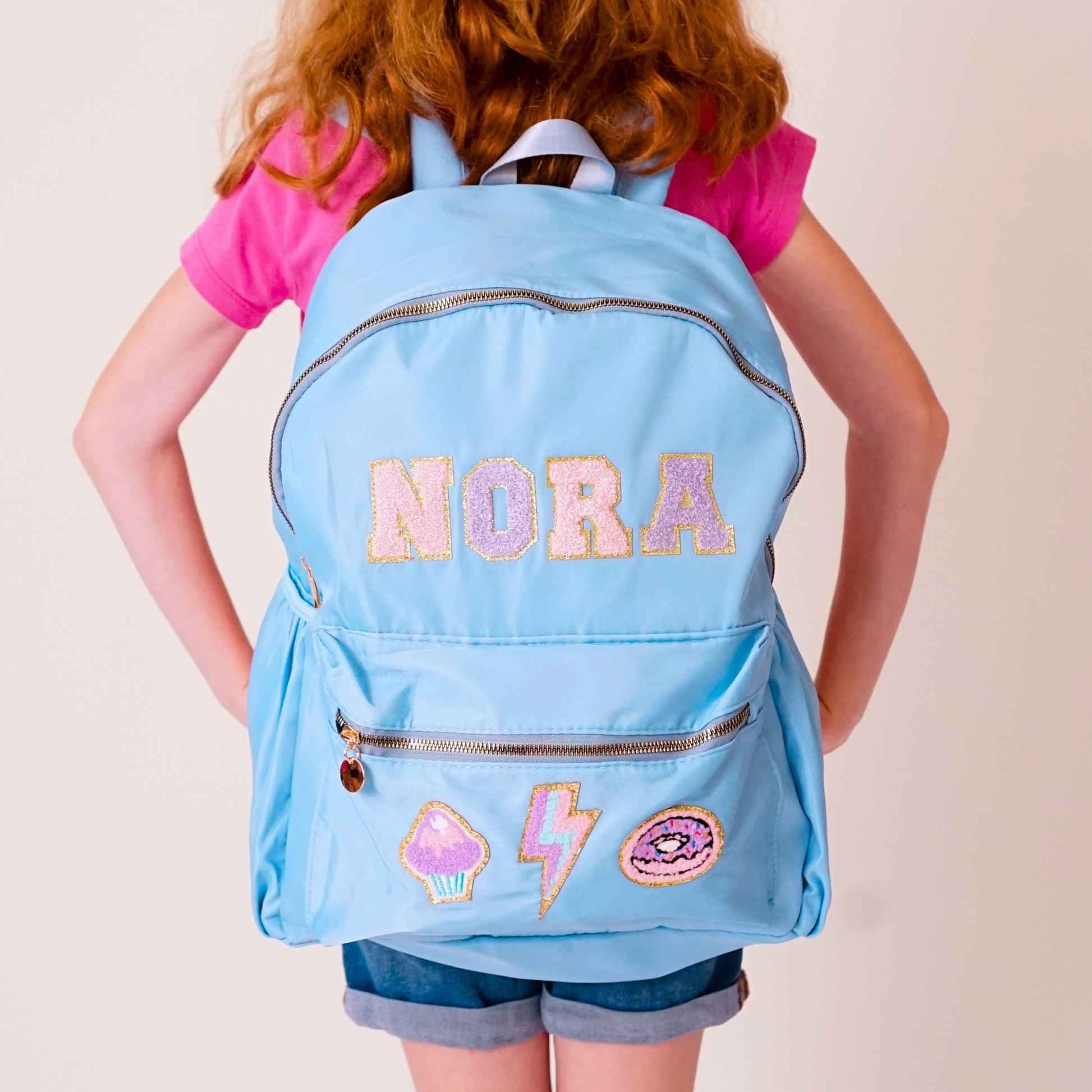 Child wearing a light blue backpack with 'NORA' and various designs on a plain background