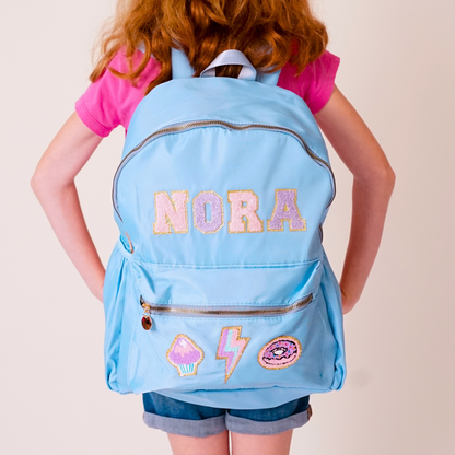 Child wearing a light blue backpack with 'NORA' and various designs on a plain background