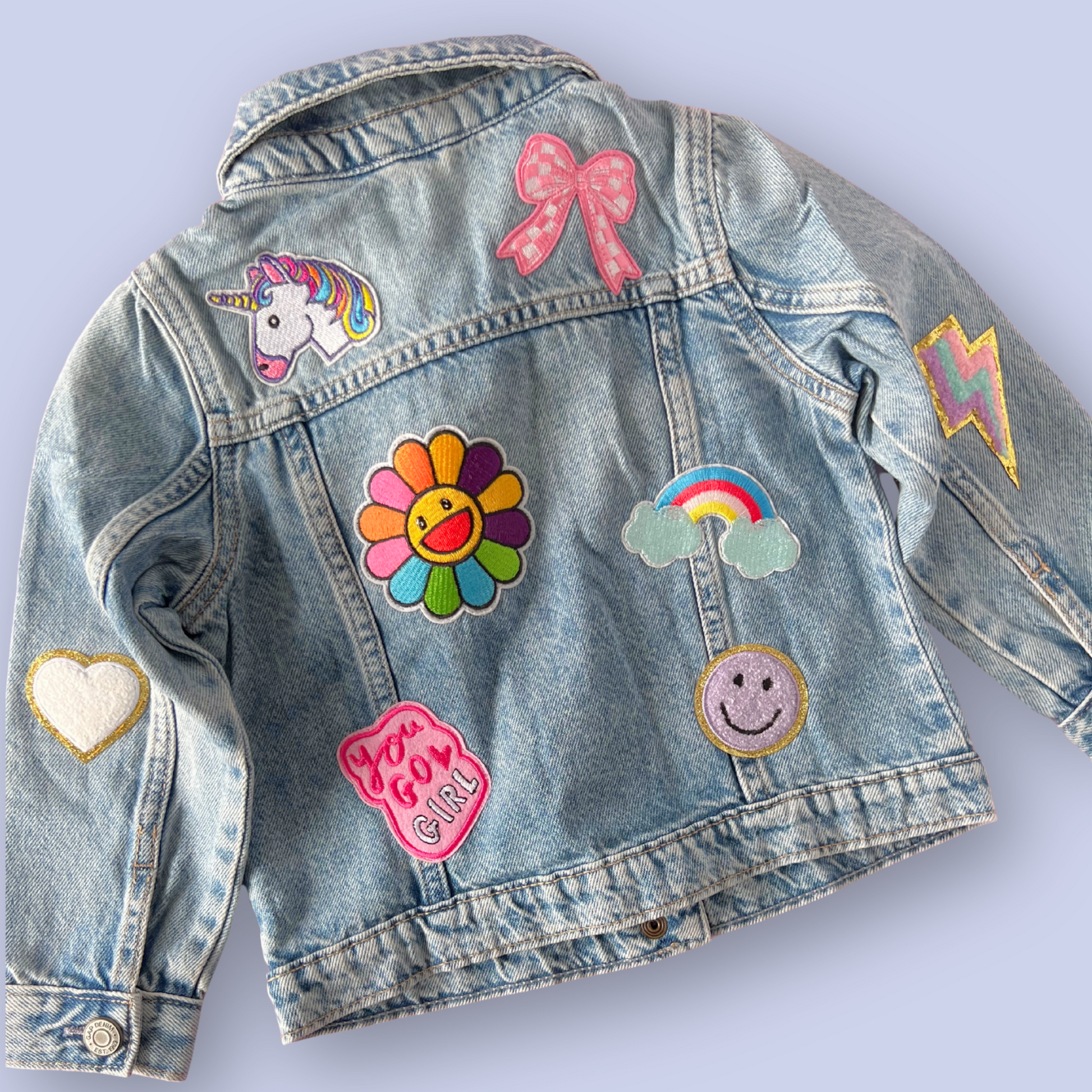 Denim jacket with colorful patches including a unicorn, flower, rainbow, and heart on a light purple background.