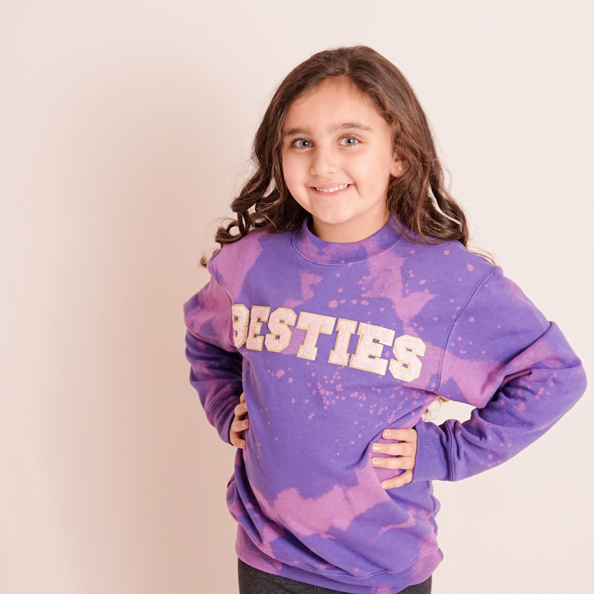 Young girl wearing a purple 'BESTIES' sweatshirt against a beige background