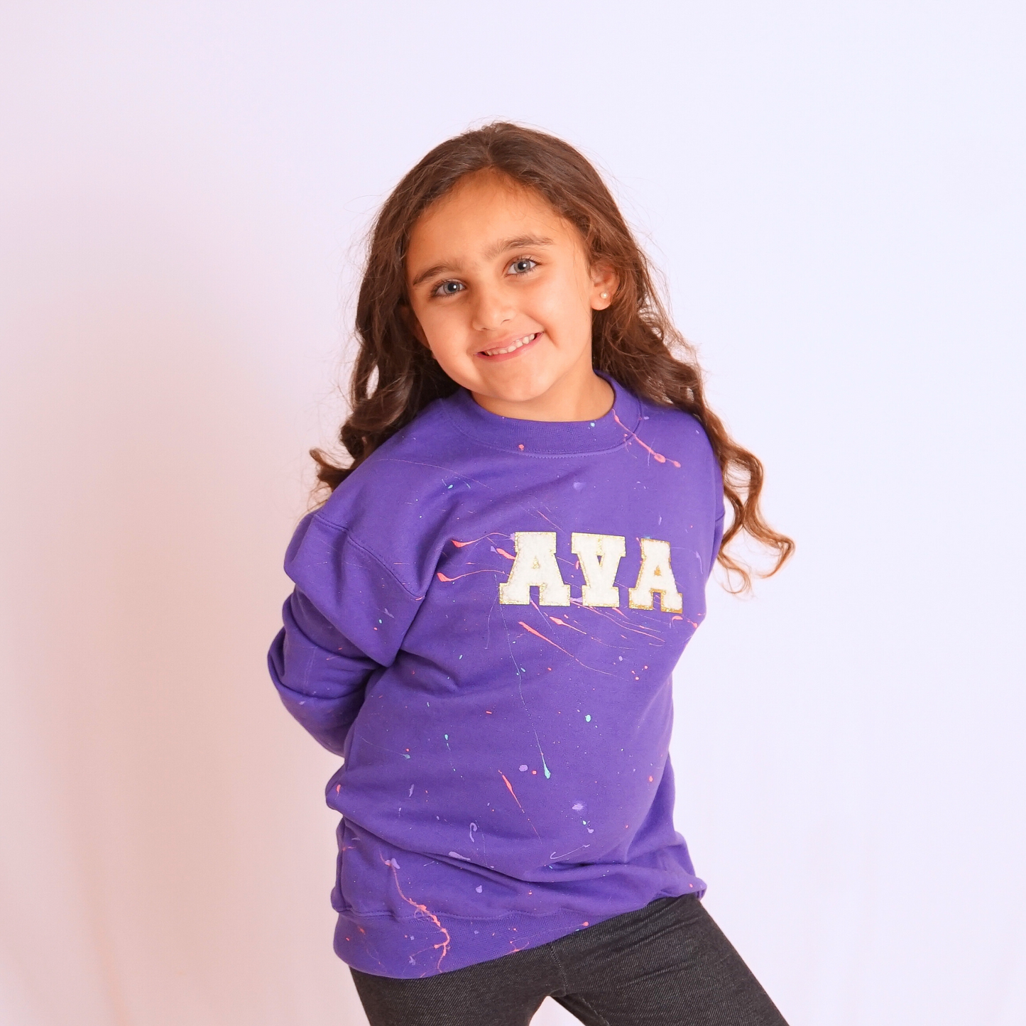 Child wearing a purple sweatshirt with 'AVA' on it against a light pink background