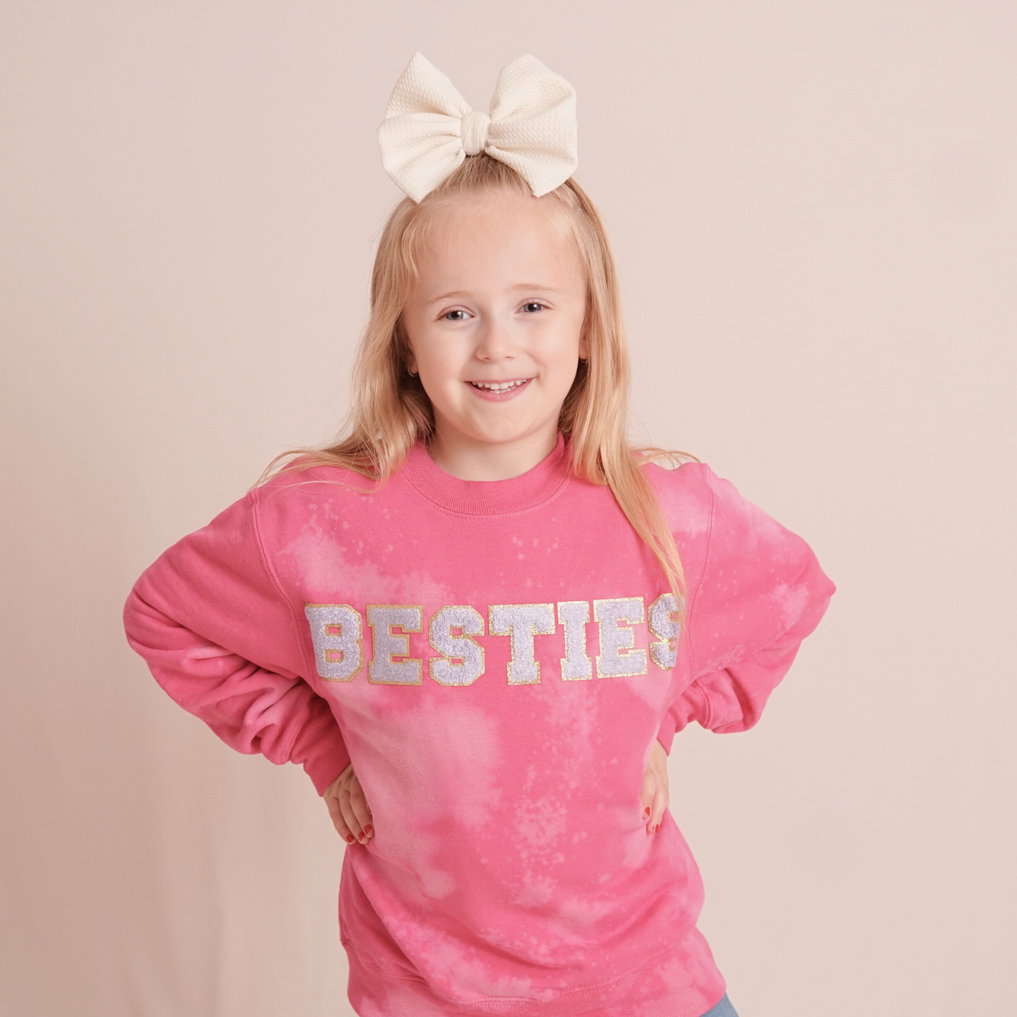 Young girl wearing a pink 'BESTIES' sweatshirt with a white bow in her hair on a beige background