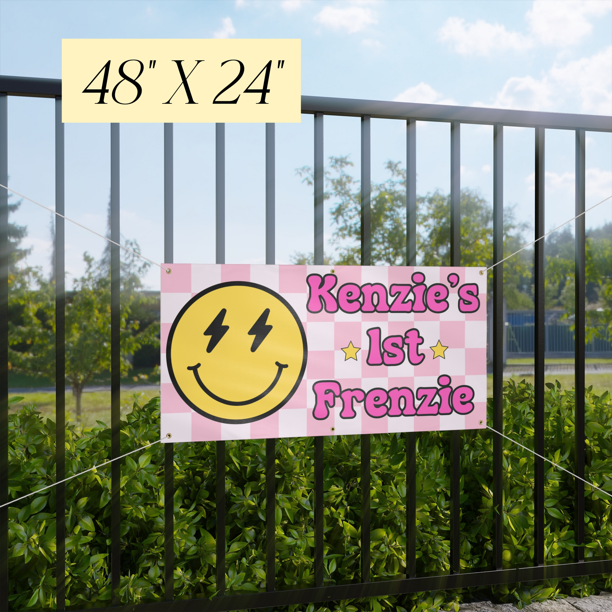 Pink checkered banner with smiley face and text 'Kenzie's 1st Frenzie' attached to a metal fence outdoors.