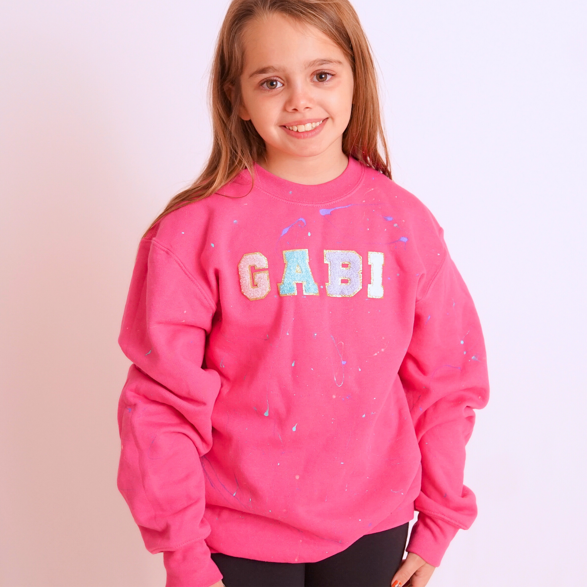 Young girl wearing a pink sweatshirt with 'GABI' printed on it against a light pink background