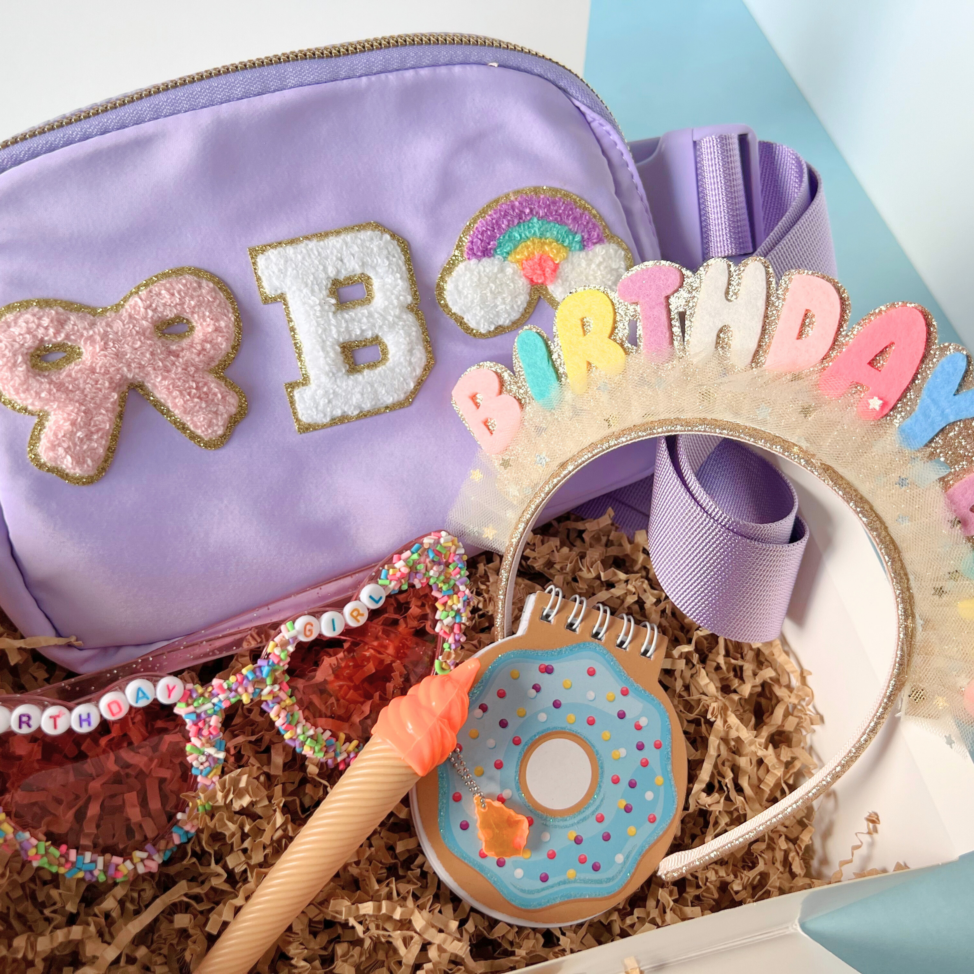 Lavender set – Light purple fanny pack with a bow,  “B” initial, surrounded by full birthday box contents in coordinating colors.