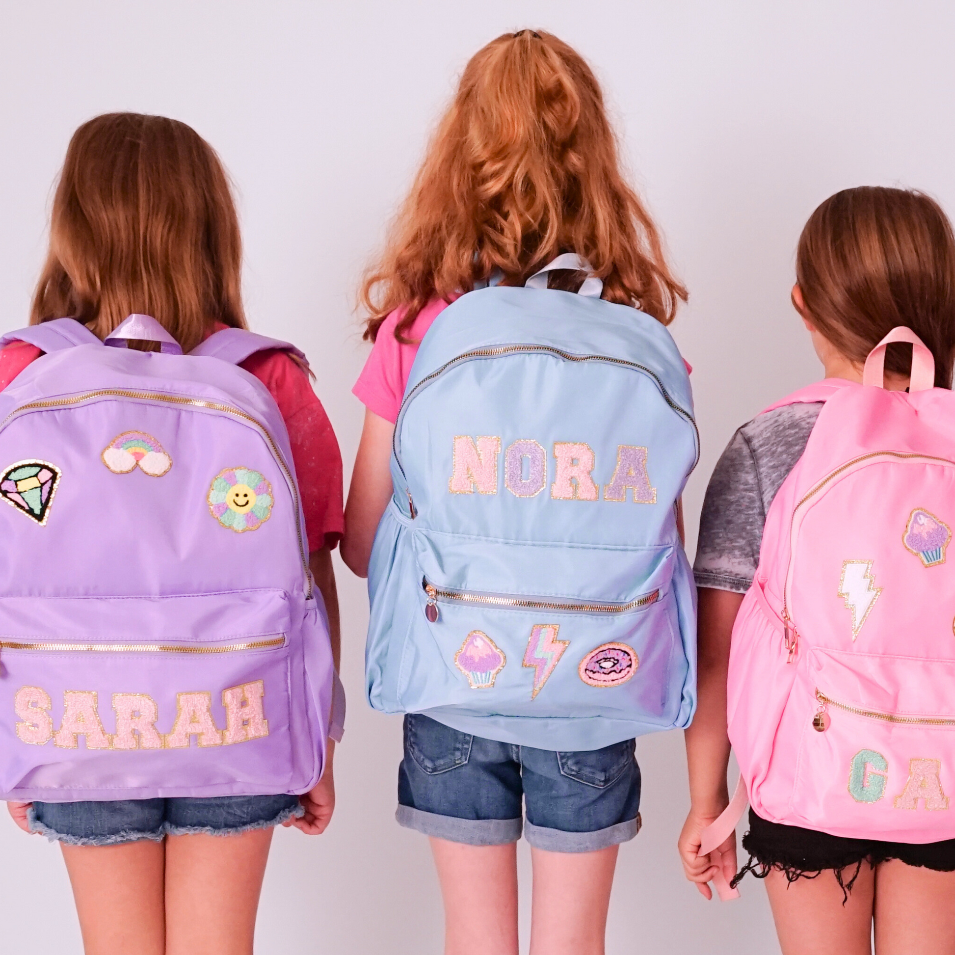 Three children wearing personalized backpacks with names on a plain background