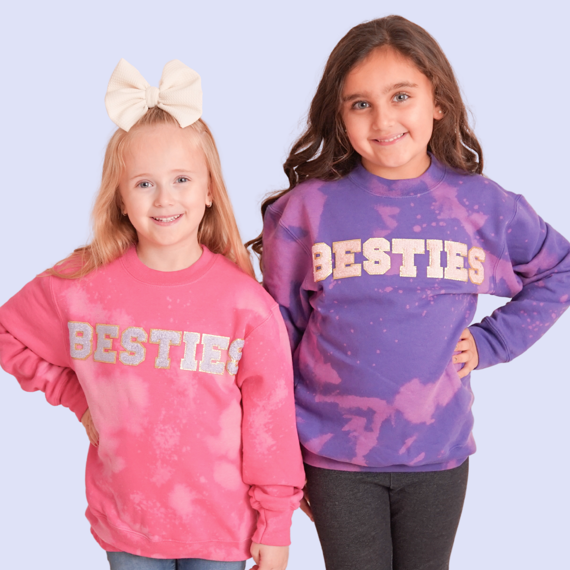 Two girls wearing “Besties” sweatshirts – one in pink bleach-dye, one in purple bleach-dye – smiling at the camera