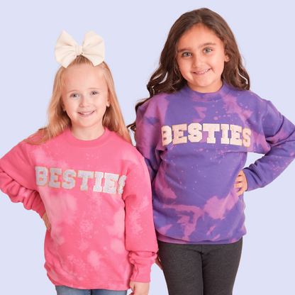 Two girls wearing “Besties” sweatshirts – one in pink bleach-dye, one in purple bleach-dye – smiling at the camera