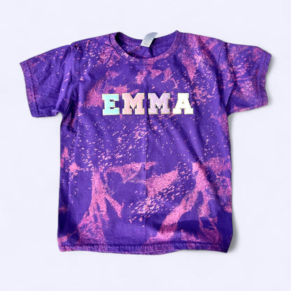 
A small-sized T-shirt, dyed in a vibrant purple and pink bleach-splatter pattern, lying flat on a solid background. The name "EMMA" is displayed prominently on the front in light-colored, pastel-textured letters.