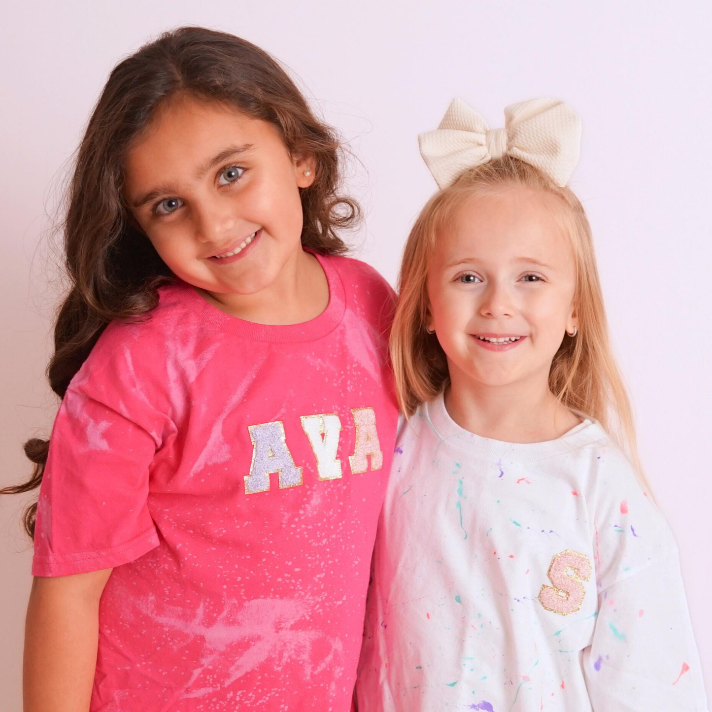 
A photograph of two young girls smiling and posing together against a plain white background. The girl on the left has long, dark, wavy hair and is wearing a hot pink T-shirt with a bleach-splatter pattern and the name "AVA" in textured, pastel-colored letters on the front. The girl on the right has long blonde hair and is wearing a large, cream-colored bow on top of her head. She is wearing a white, paint-splattered T-shirt with a single textured, embroidered letter 'S' on the front.
