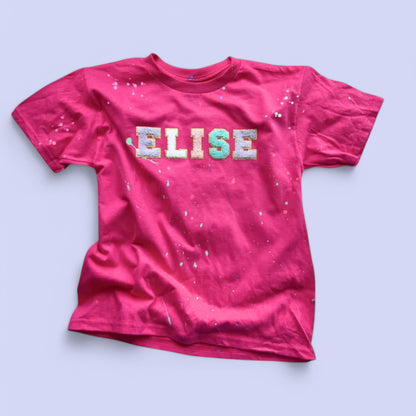 A pink custom name t-shirt with the name 'ELISE' written across the front in multicolored letters, featuring a paint splattered design.