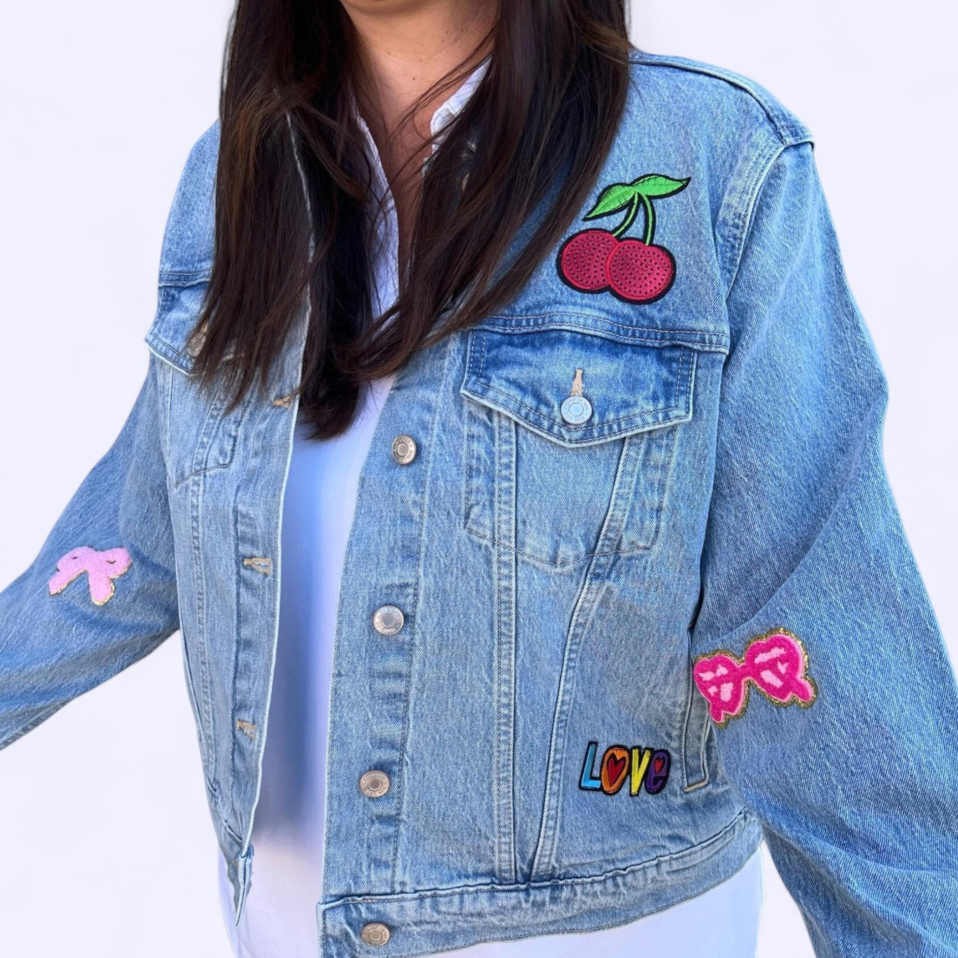 
A cropped, light-wash denim jacket adorned with various patches. On the left chest pocket is a colorful "LOVE" patch. A large, red cherry patch with a green stem is on the upper right side. The right sleeve has a large pink bow patch, and the left sleeve has a smaller, similar pink bow patch. The person wearing the jacket is not fully visible, with the focus on the jacket and its unique patches.