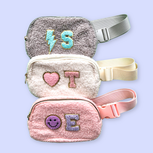 Gray, cream, and pink sherpa fanny packs, each with colorful chenille letter patches and glitter edges, displayed on a clean pastel background.