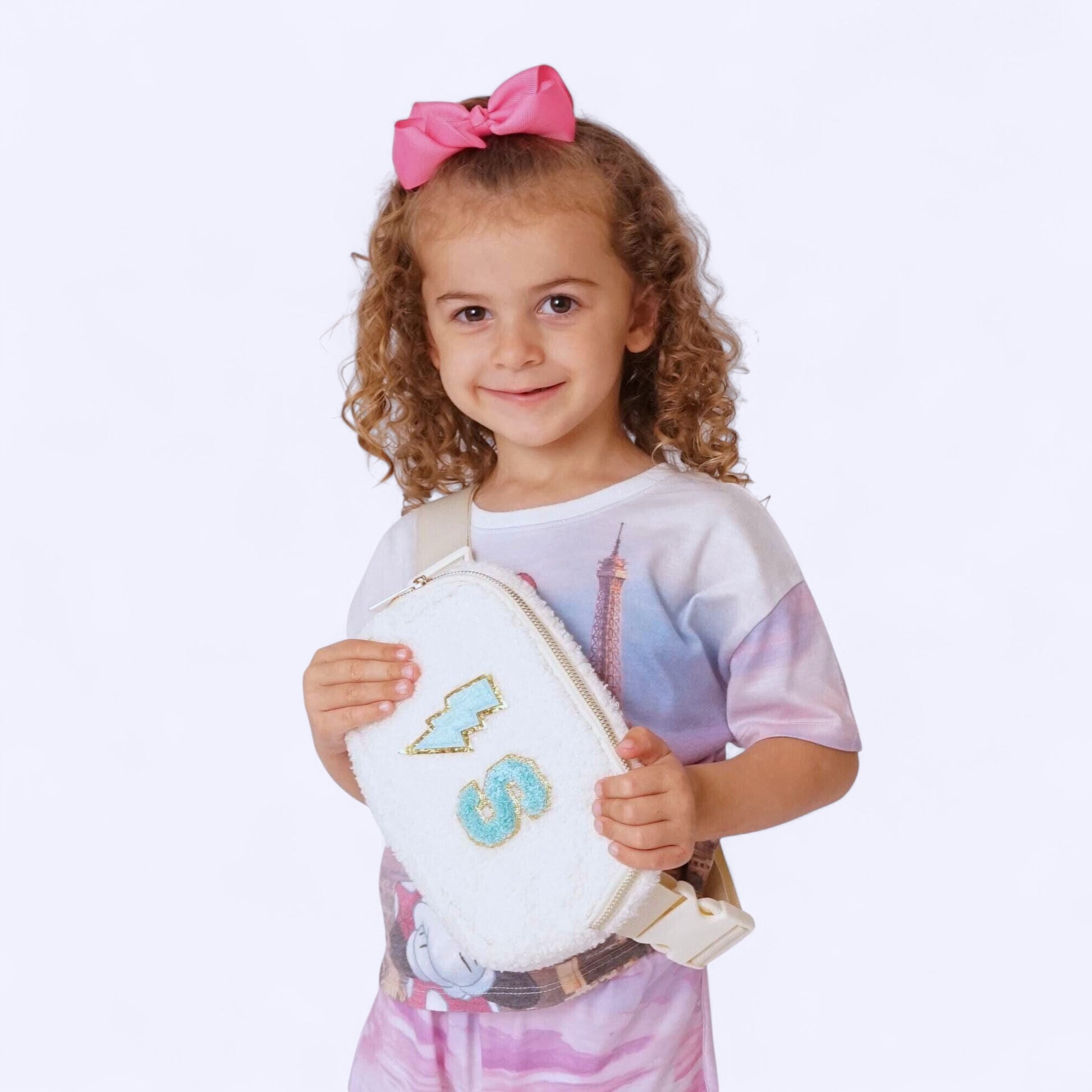 A smiling girl wearing a white tee and pink skirt holds a white sherpa fanny pack with pastel chenille letters and a lightning bolt patch